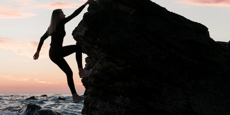 Woman Climbing Jugged Rock Next To Ocean