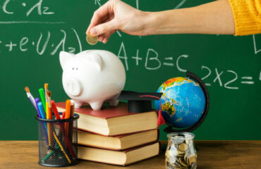 Hand Putting Coin In Piggy Bank Surrounded By Books, Pens, A Globe and A Jar of Coins