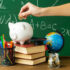 Hand Putting Coin In Piggy Bank Surrounded By Books, Pens, A Globe and A Jar of Coins