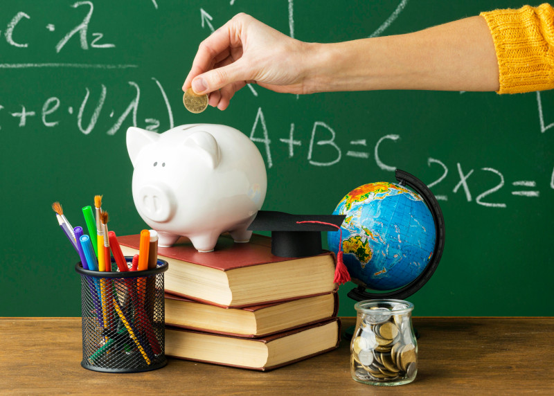 Hand Putting Coin In Piggy Bank Surrounded By Books, Pens, A Globe and A Jar of Coins