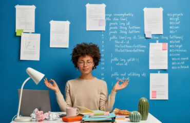 Woman meditating at a desk with laptop, crumpled pieces of paper, a cup of coffee and blue board in the background with posted notes and pseudocode
