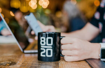 Woman hands sitting n front of laptop holding a mobile phone and a mug with the 80/20 Written on it