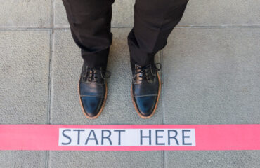 Man's lower section of legs wearing formal shoes standing in from of a starting line with the text "Start Here"