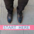 Man's lower section of legs wearing formal shoes standing in from of a starting line with the text "Start Here"