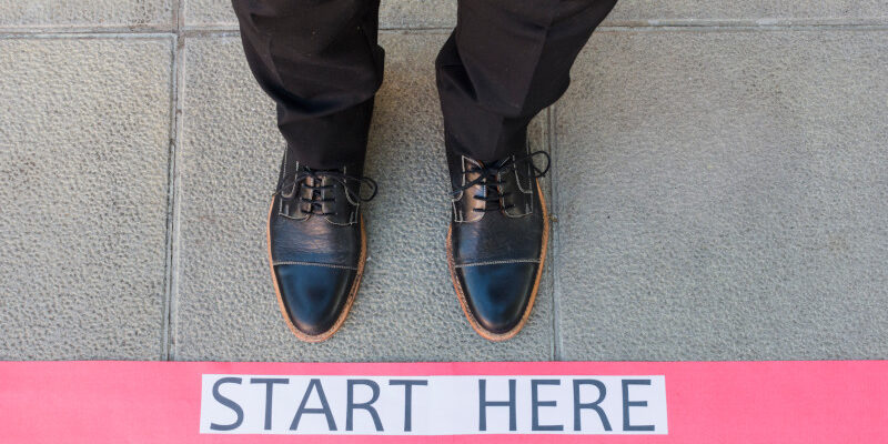 Man's lower section of legs wearing formal shoes standing in from of a starting line with the text "Start Here"