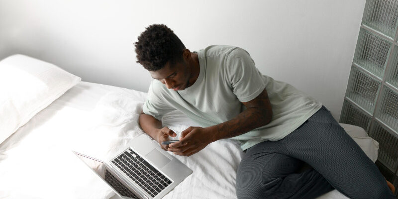 Young man looking at phone whilst lying on be with laptop on it