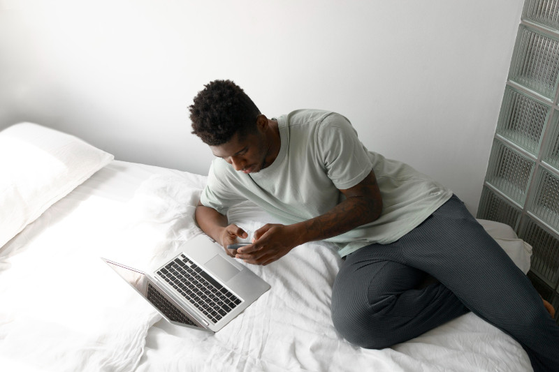 Young man looking at phone whilst lying on be with laptop on it