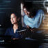 Woman working with code and man looking over her shoulder whilst holding a tablet all with code and chart overlays