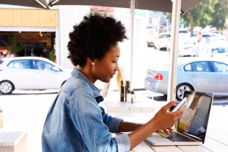 Woman using cellphone with laptop outside cafe