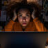 A woman in a dark room illuminated by a single lightbulb anxiously looking at her laptop whilst holding her head